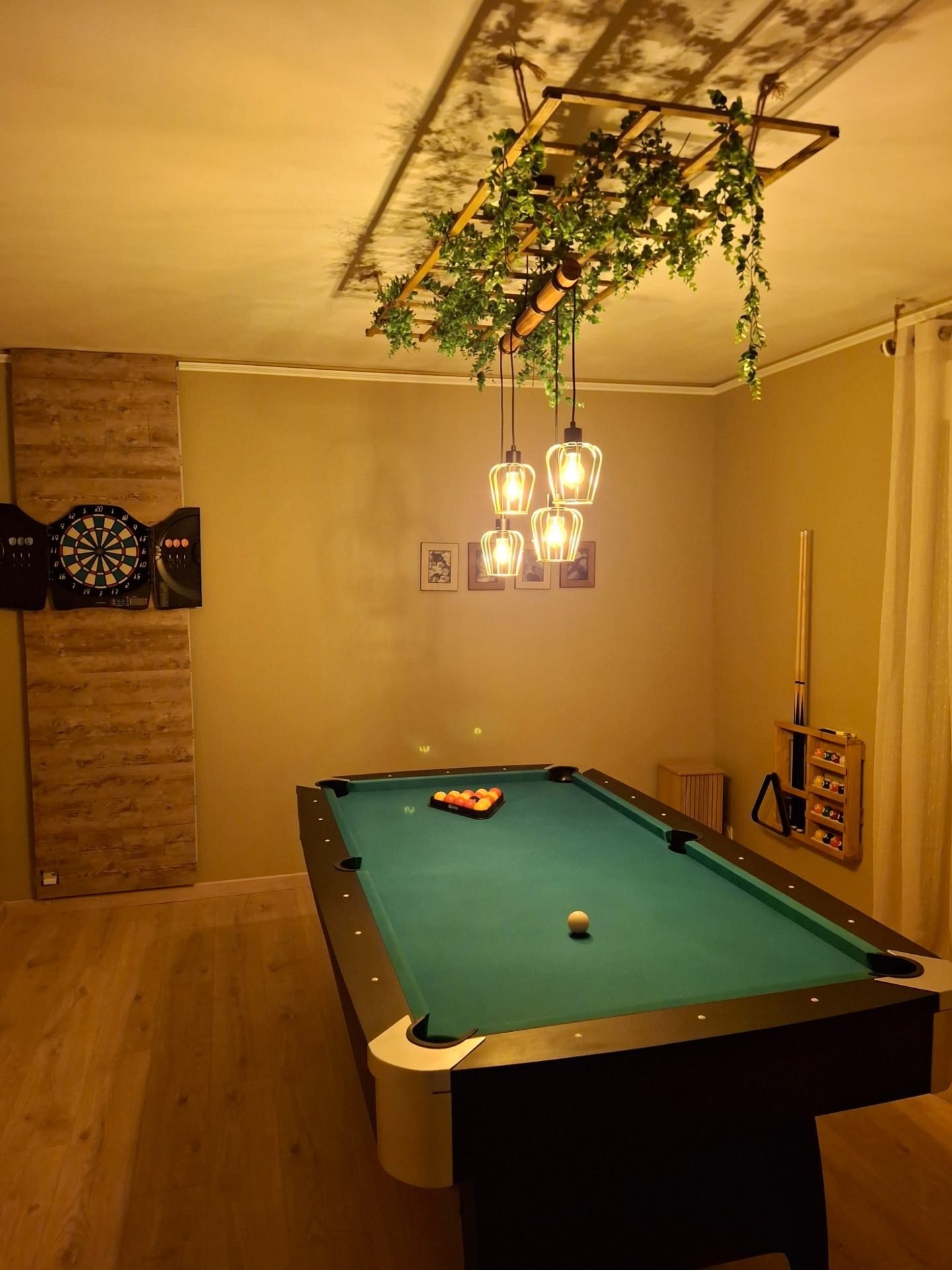 Salle de billard : table au tapis vert sous des suspensions dorées et une treille de feuillages, jeu de fléchettes au mur