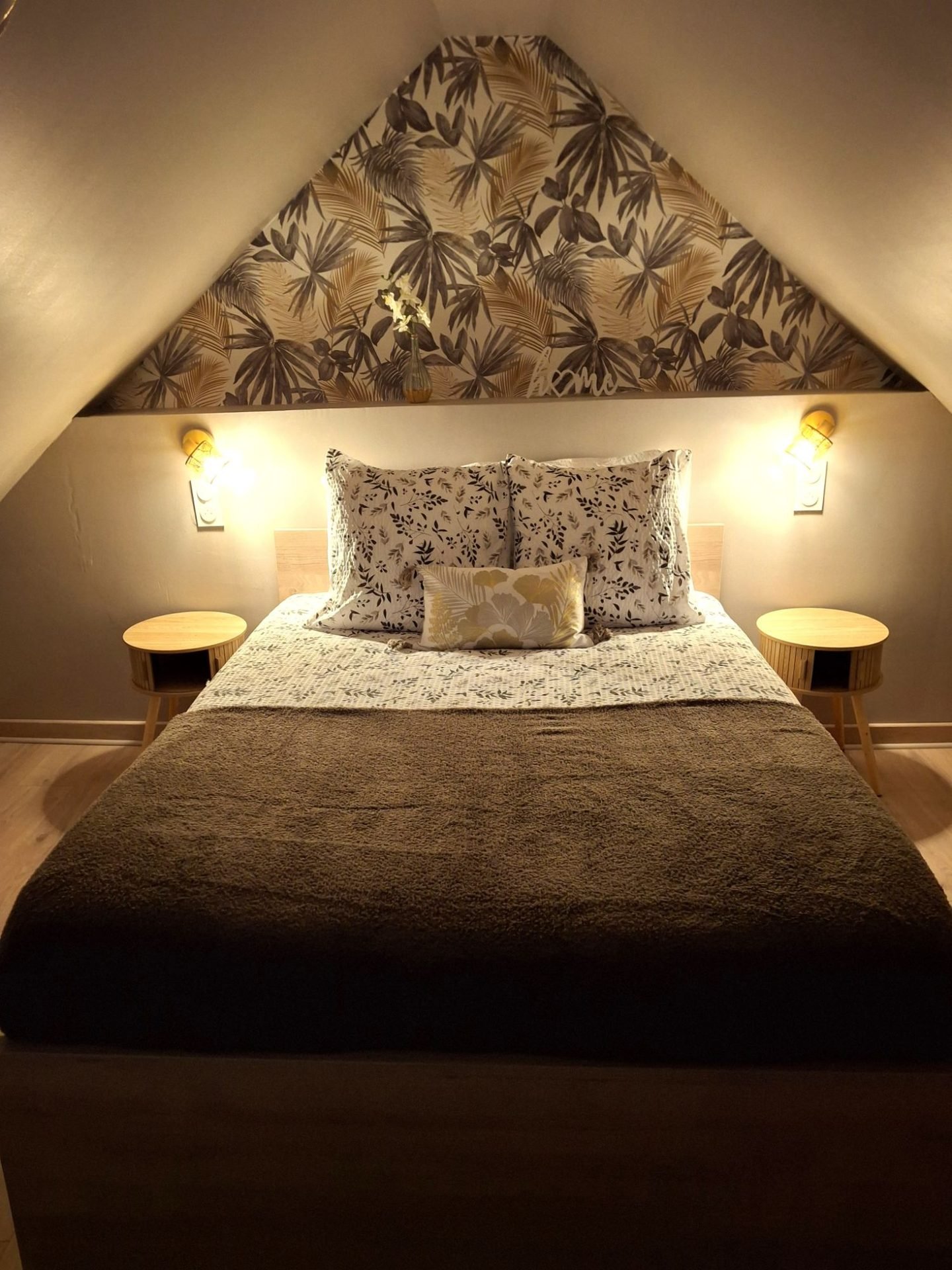 Chambre sous les combles : lit double, papier peint de palmes dorées, deux appliques et tables de chevet rondes en bois