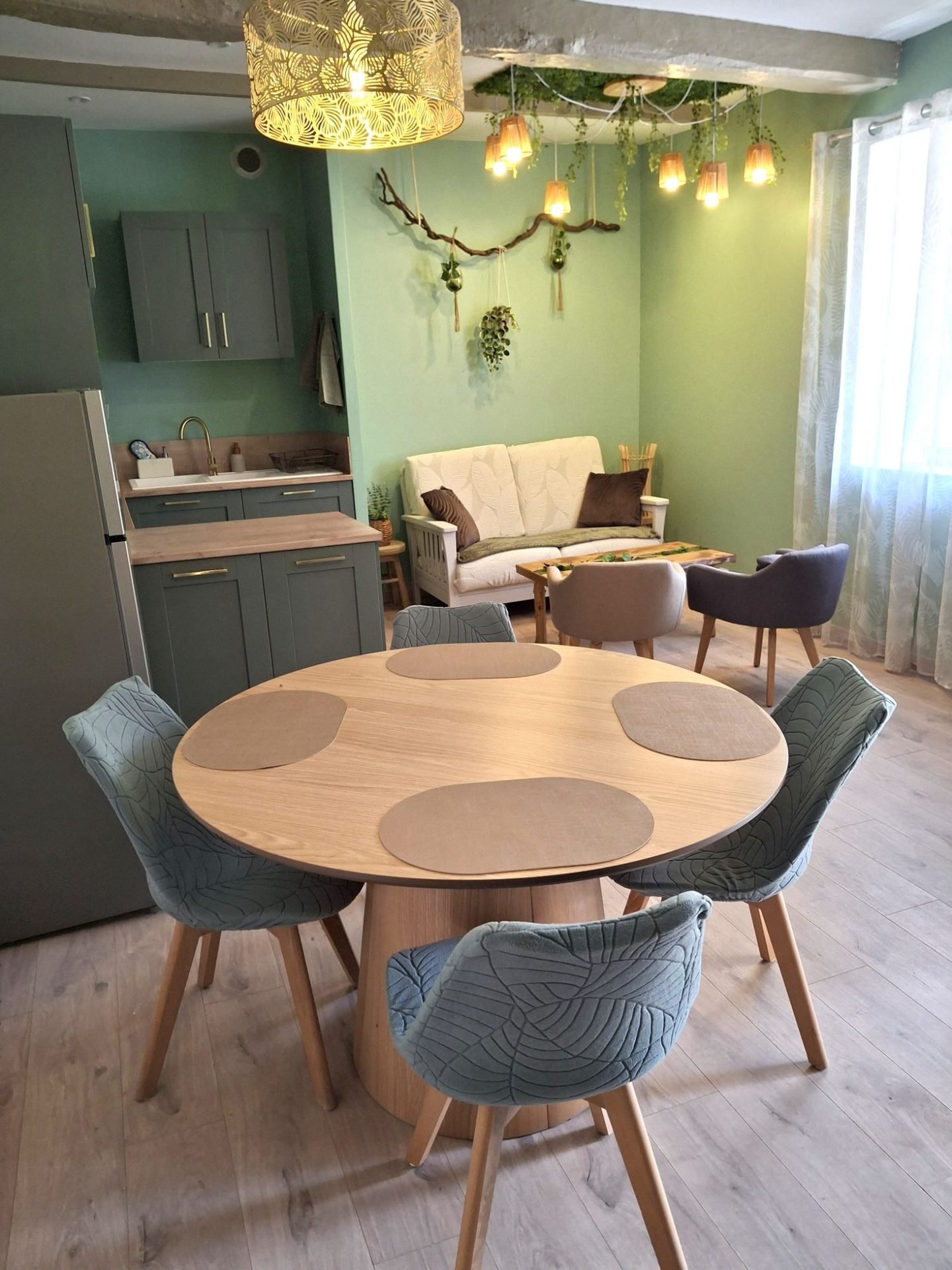 Table ronde en bois et quatre chaises aux motifs de feuilles, cuisine vert sauge, canapé et suspensions végétales au plafond