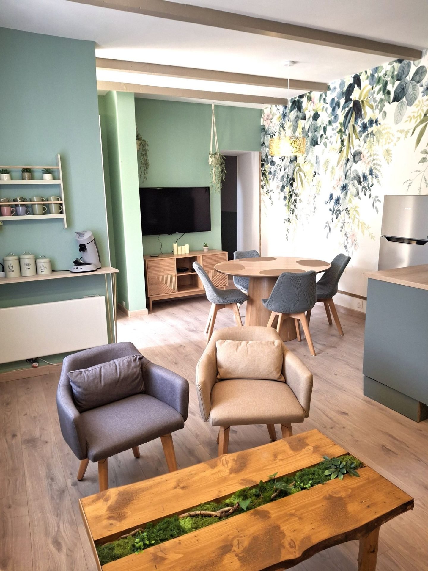 Pièce de vie : deux fauteuils devant une table basse en bois garnie de mousse, coin café, télévision, table ronde et papier peint de feuillages
