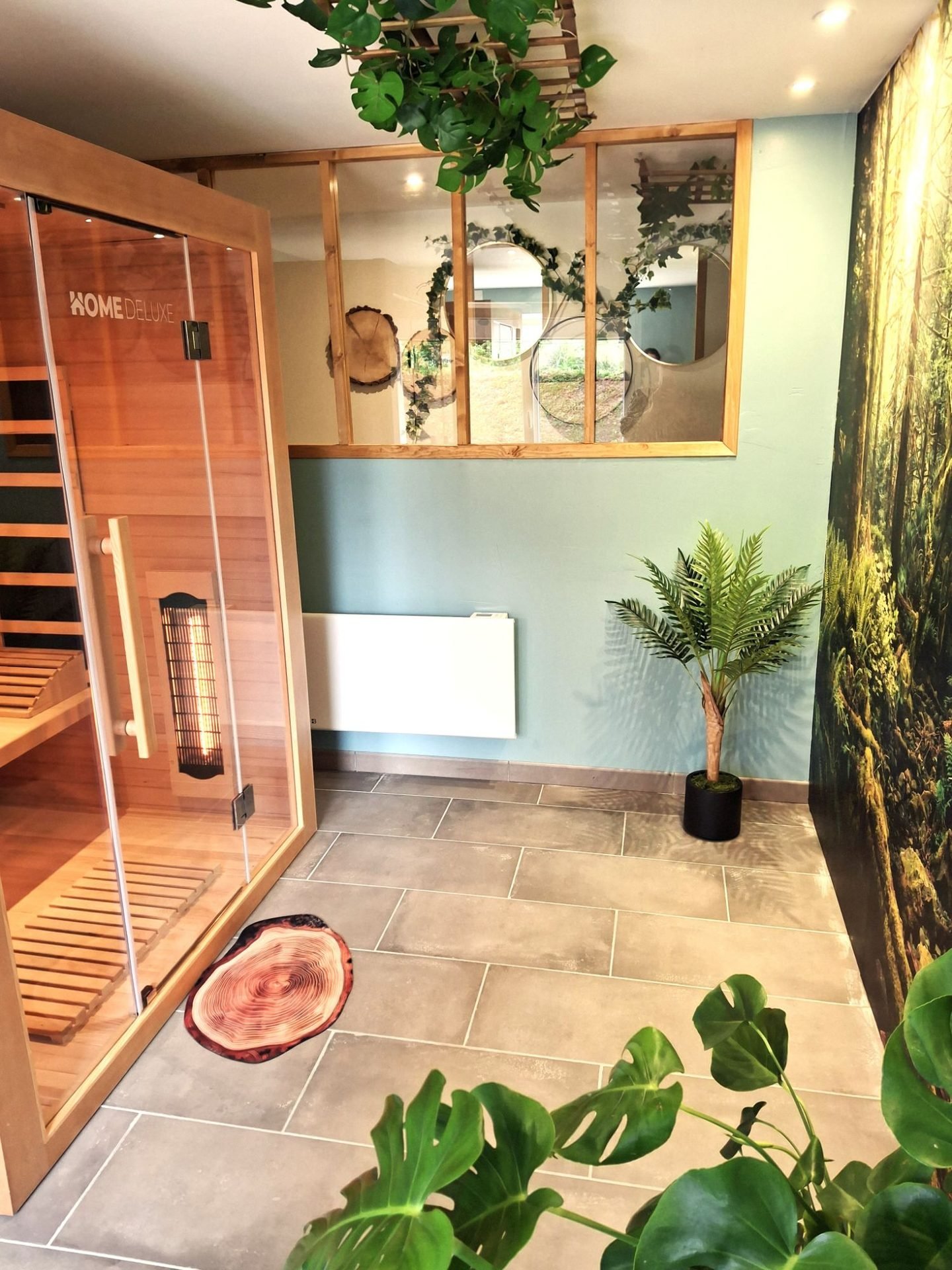 Espace sauna : cabine en bois vitrée, carrelage gris, plantes et grand décor de forêt au mur