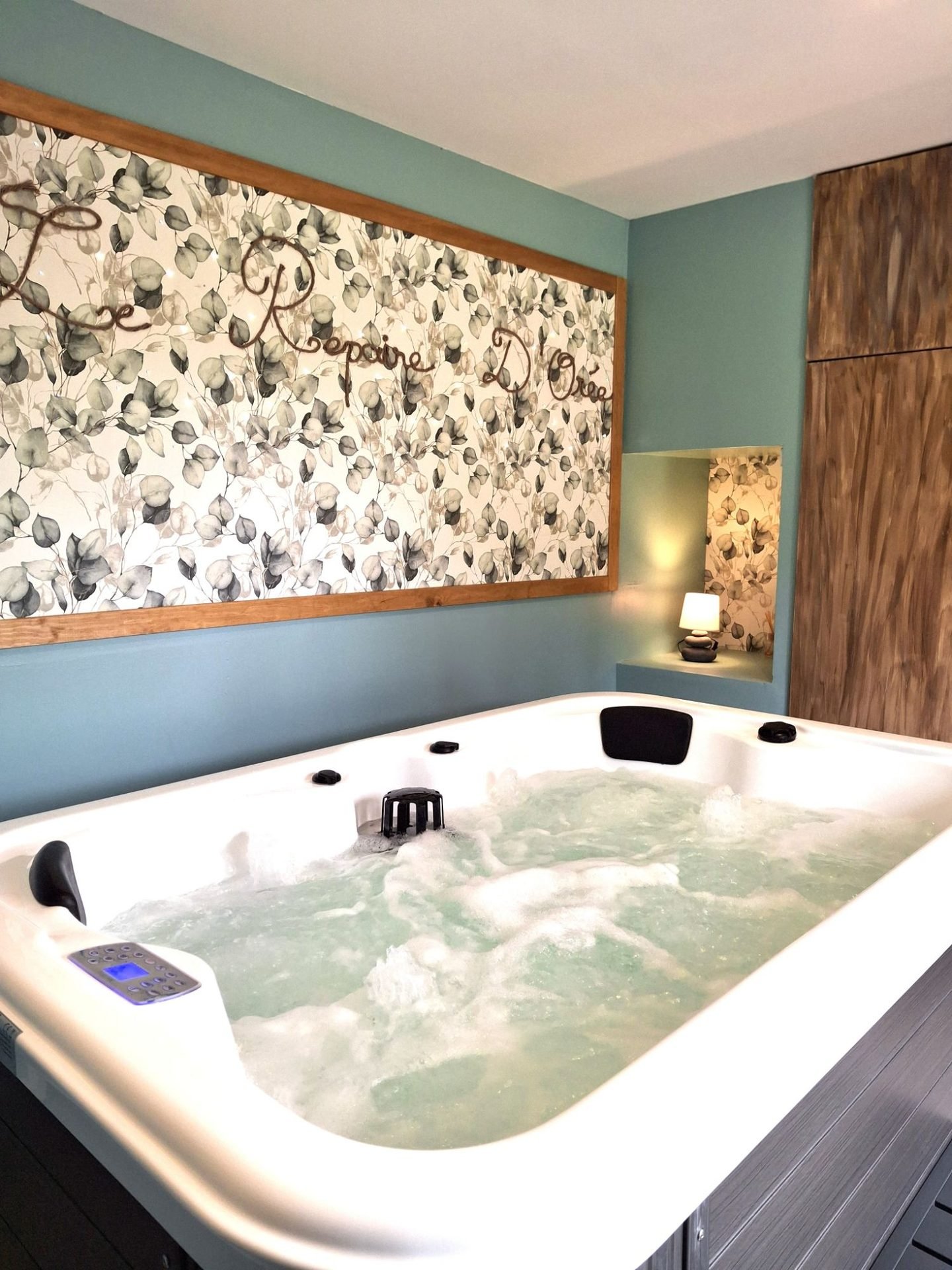 Le jacuzzi devant le grand mur aux feuilles d'eucalyptus où est écrit « Le Repaire d'Orée », une niche éclairée par une petite lampe